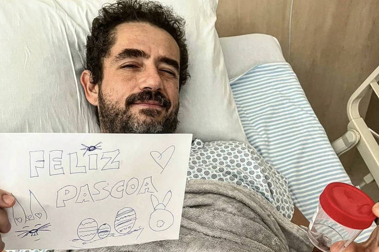 Homem branco de cabelos pretos está deitado em cama de hospital. Ele segura um papel com desenhos e o escrito "Feliz Páscoa" 