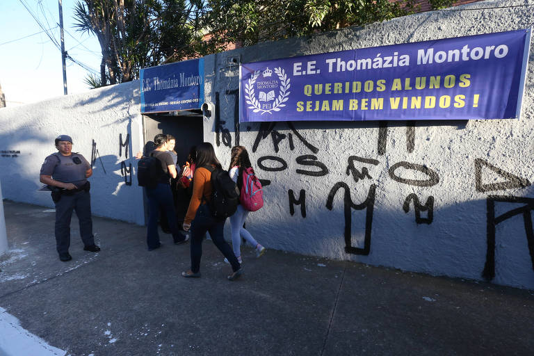 Estudantes de mochila na frente da escola; no muro, há um cartaz com a mensagem: "Queridos alunos,  sejam bem-vindos!"
