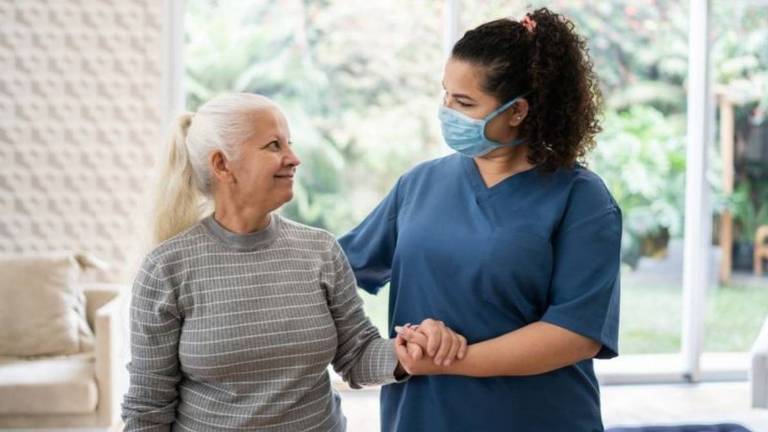 Enfermeira, de máscara, segura e olha para mulher idosa