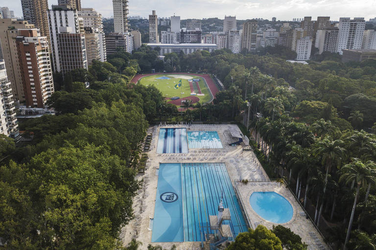 Visão aérea do Clube Pinheiros, na zona oeste de São Paulo, com três piscinas em primeiro plano e um campo de futebol ao fundo, entre muitas árvores