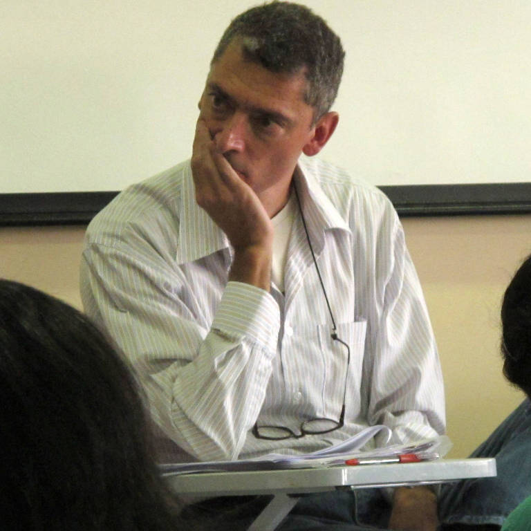 Homem com camisa branca põe a mão no queixo e olha compenetrado em sala de aula