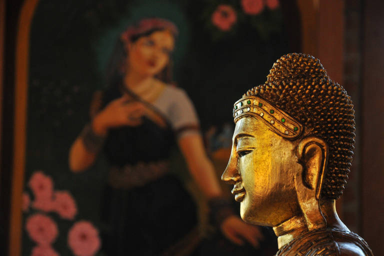 Imagem de uma estátua dourada que simboliza o Buda. Ao fundo, um quadro de uma mulher com a mão no peito e flores rodeando