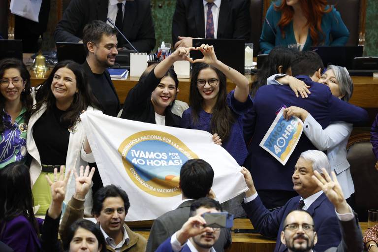 Foto mostra duas deputadas chilenas brancas com cabelos pretos lisos juntas formando um coração com as mãos e segurando uma bandeira que diz "Vamos pelas 40 horas", no centro da Câmara dos Deputados do país, cercadas por outros parlamentares. Elas comemoram a aprovação da redução das horas de trabalho no Chile