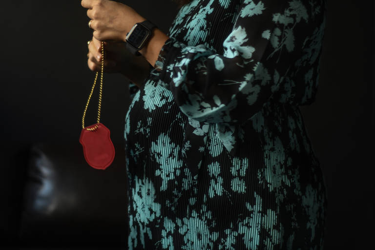 Imagem colorida mostra a barriga de uma mulher grávida, voltada para o lado esquerdo e segurando o distintivo da Polícia Civil. Ela usa um vestido com desenhos nas cores preta e verde.
