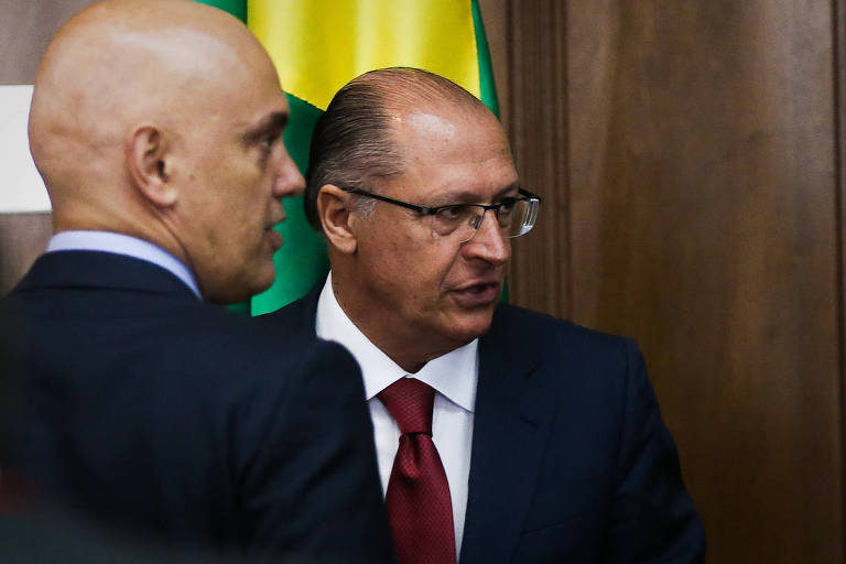 Alexandre de Moraes está de costas, no canto esquerdo da foto, olhando para a direita, e Alckmin está a sua frente, olhando na mesma direção. Os dois são brancos e vestem camisa clara e paletó escuro. Estão em ambiente fechado com bandeira do Brasil e parede de madeira ao fundo