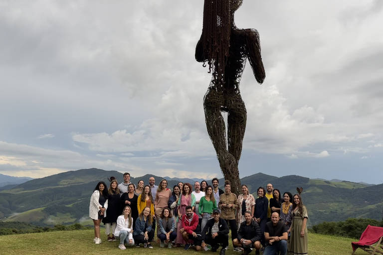 Cerca de trinta participantes do encontro se reúnem em frente à escultura de ferro com nove metros de altura, posicionada no alto da serra. Ao fundo, demais montanhas da Mata Atlântica mineira compõem o cenário.