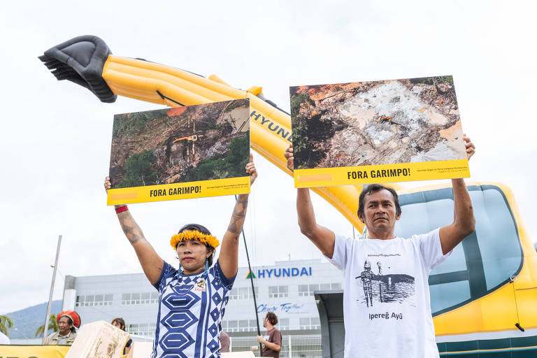 Mulher e homem indígenas seguram cartazes com fotos de áreas de garimpo em frente a uma retroescavadeira amarela inflável; ao fundo é possível ver o prédio da Hyundai, com o logotipo da empresa