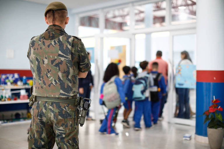 Homem com farda camuflada e boina verde-oliva, de costas e com pistola no coldre, observa crianças dentro de escola