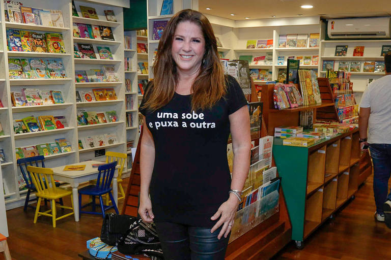 Em foto colorida, mulher de camisa preto posa para foto em uma livraria