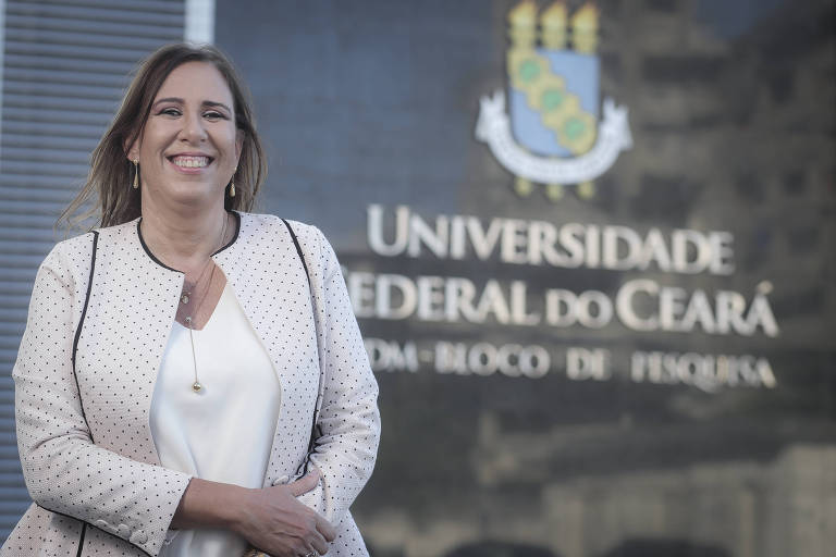 Mulher branca, de roupa clara, posa sorridente em frente à entrada da Universidade Federal do Ceará