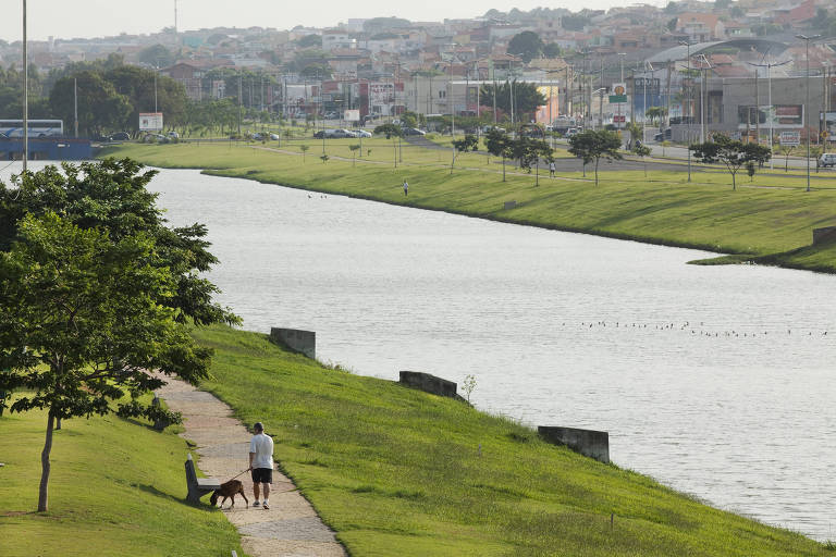 Foto mostra rio e parque. É possível ver um homem passeando com um cachorro. Ao fundo, há casas.