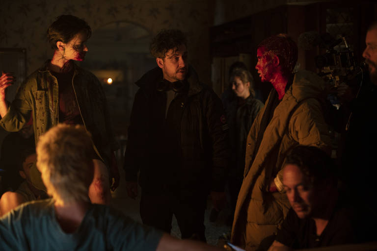 Em set de filmagens à meia-luz, um homem instrui dois atores em cena, ambos cobertos de sangue e vestindo casacos. Ao redor, a equipe técnica