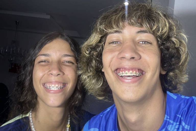 Duas pessoas aparecem na foto, lado a lado. Na esquerda, uma menina usando camisa da CBF azul marinho e colares sorri, mostrando os dentes com aparelho. Na esquerda, menino de cabelos loiros usa camisa de futebol azul e sorri, também mostrando o aparelho dental. Eles são muito parecidos.