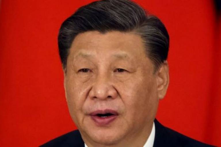 Xi Jinping é um homem asiático, de cabelos lisos e curtos e sem barba. Veste terno preto e camisa branca. Ao fundo da imagem, há um pano vermelho