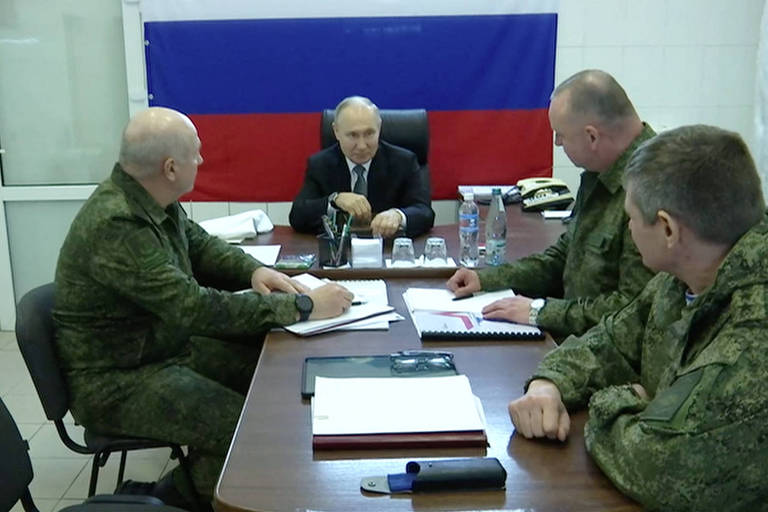 Putin sentado à mesa com três homens em uniformes camuflados; ao fundo, bandeira da Rússia