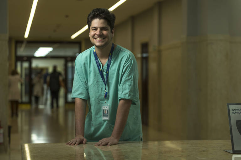 Imagem colorida mostra Fernando Brito, um homem branco de 26 anos; ele veste um uniforme médico na cor verde, está com as duas mãos apoiadas em uma mesa e sorri.