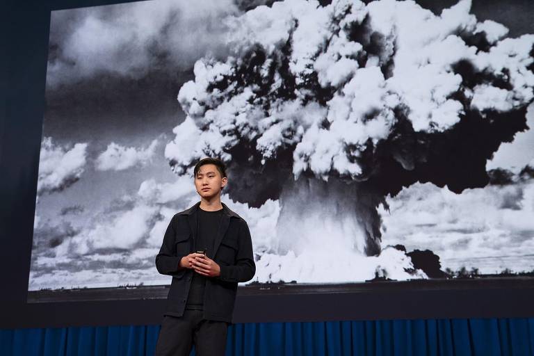 Alexandr Wang é um jovem amarelo, de cabelos pretos lisos. Ele veste blazer sobre camiseta preta e está à frente da foto do cogumelo de fumaça de uma bomba atômica.