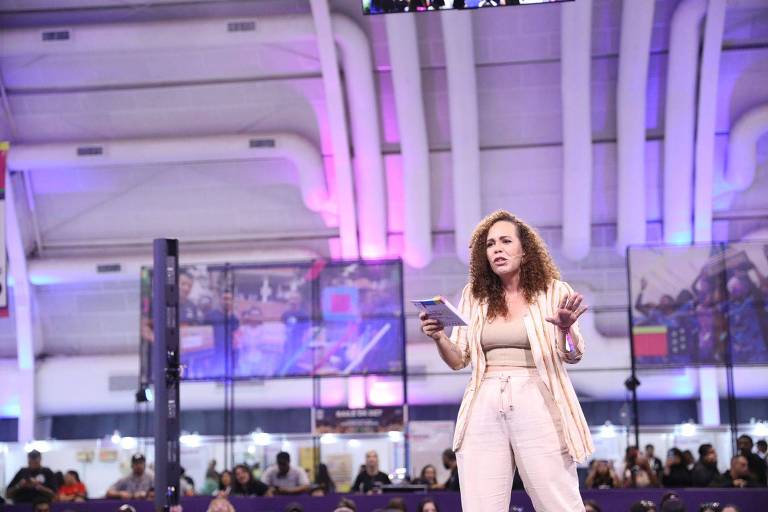 Kalyne Lima, mulher não-branca, de cabelos cacheados; está em pé em um palco e fala para o público; ela usa roupas de cor rosa claro