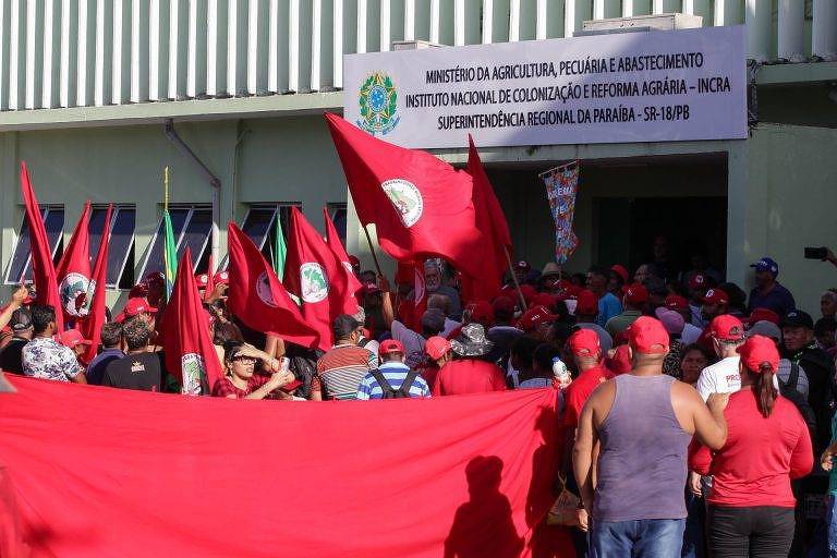 Grupo de manifestantes ergue bandeira horizontal vermelha. Ao funo placa sinaliza entrada da sede do Incra