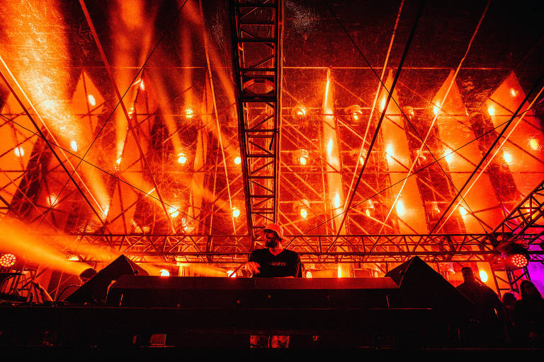 DJ se apresenta para plateia; ao fundo, várias luzes avermelhadas se sobrepõe à noite escura