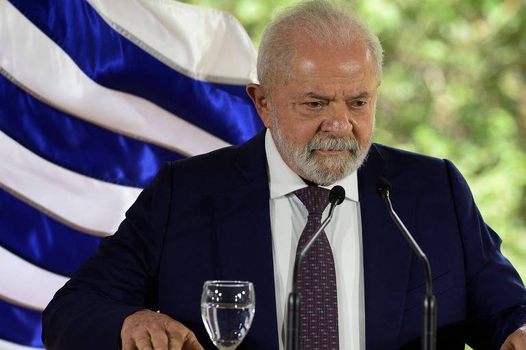 O presidente Lula, de terno escuro e gravata azul, fala em frente a um microfone, em visita ao Uruguai; ele está de pé, com uma taça com água perto de sua mão direita e a bandeira de listras azuis e brancas do Uruguai aparece atrás do mandatário