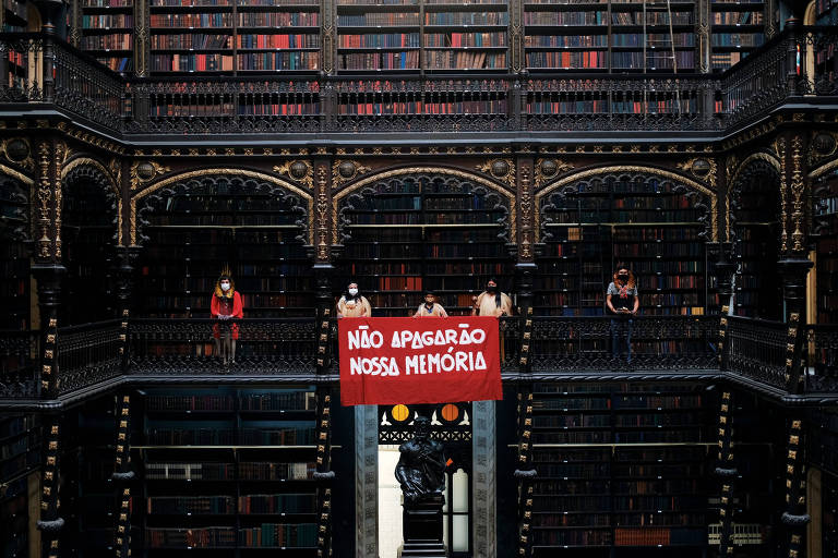 Pessoas seguram um cartaz ou pano vermelho que diz  "Não Apagarão Nossa Memória", escrito com letras brancas; elas o seguram do segundo andar da biblioteca, que é muito tradicional e antiga, com imensas estantes repletas de livros