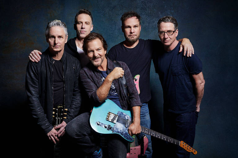 Cinco homens posam para a câmera, diante de um fundo escuro; eles vestem roupas escuras e um deles, no centro, está sentado sorrindo com uma guitarra no colo