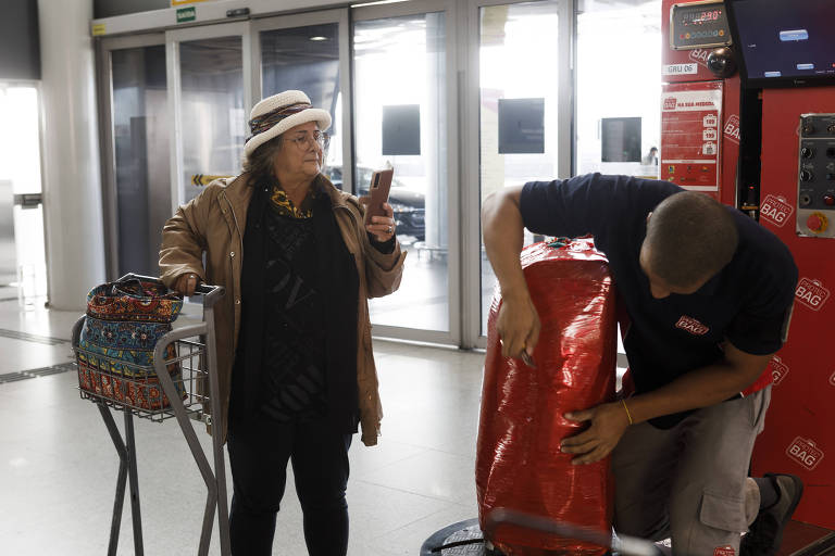 Mulher aparece em pé com celular na mão e filma homem que passa plástico em sua bagagem 