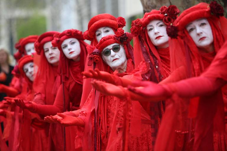 Com expressão séria, mulheres vestidas de vermelho da cabeça aos pés, apenas com o rosto aparente, pintado de branco, esticam o braço direito enfileiradas, como em uma performance teatral