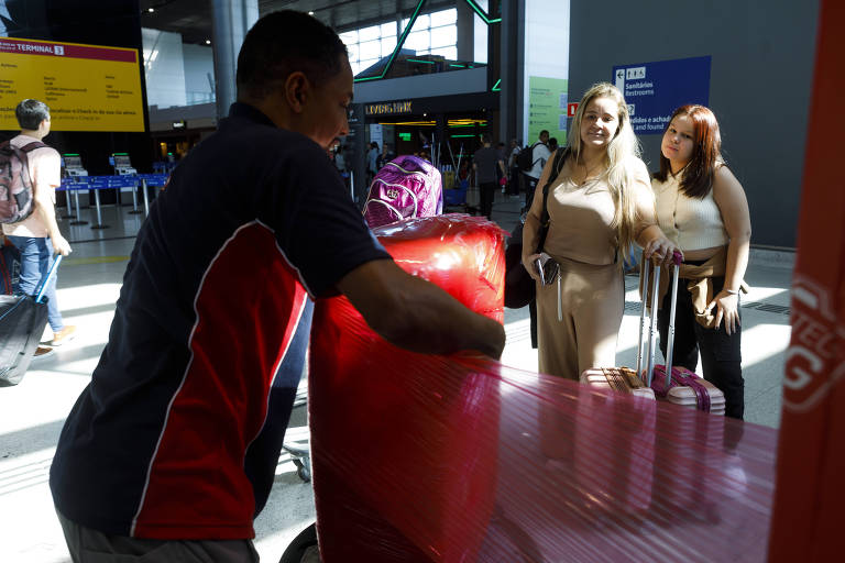 Homem à esquerda passando plástico em mala e, à direita,  duas mulheres brancas, uma mais mais velha e outra adolescente, observam a ação