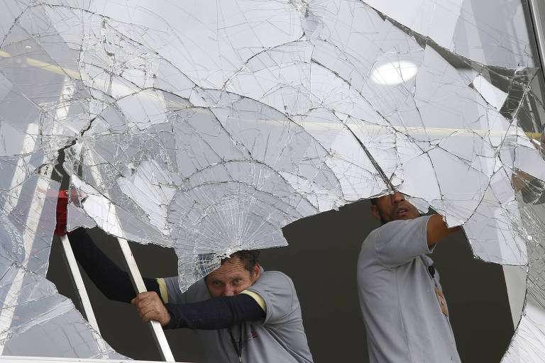 Dois homens de vestindo camisetas de cor cinza trabalham dentro de um prédio com uma grande janela quebrada
