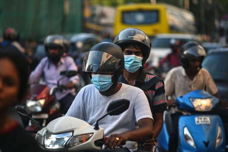 Um homem usando camiseta branca, máscara contra Covid e capacete dirige uma moto; um outro homem vai na garupa