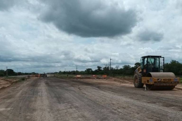 Foto mostra uma estrada sendo asfaltada no meio de um campo aberto; à esquerda está um trator
