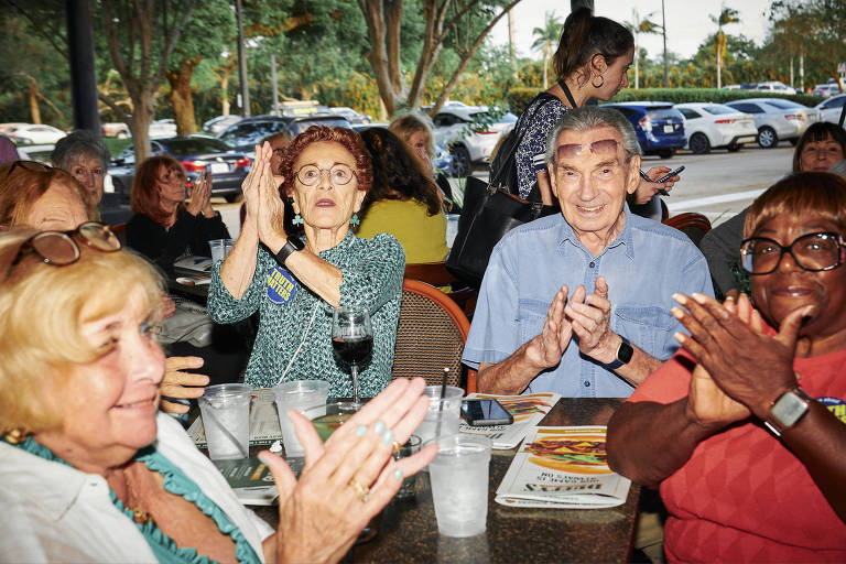 Pessoas mais velhas estão sentadas em uma mesa de restaurante, com copos e taças; elas parecem felizes, sorrindo e batendo palmas 