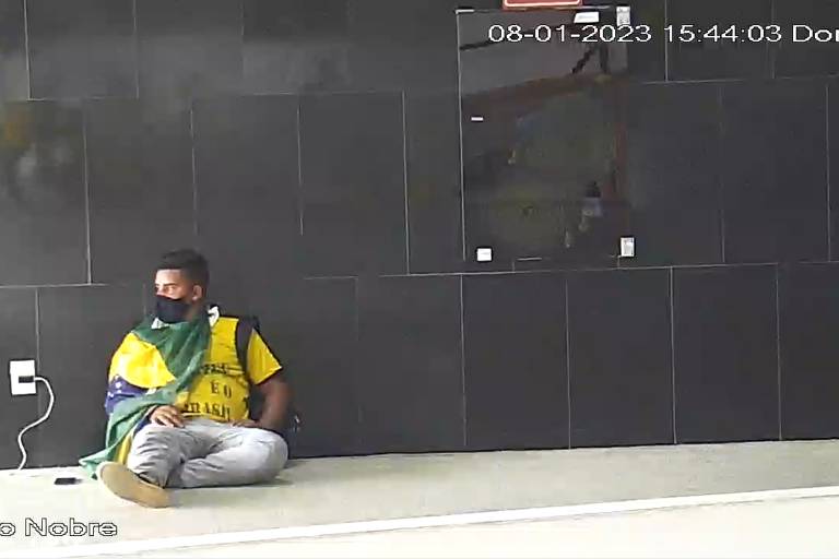 Homem carrega o celular no Palácio do Planalto durante invasão à sede dos Poderes, em 8/1. Ele estava sentado no chão e usa camiseta amarela, máscara preta e tem uma bandeira do Brasil amarrada ao corpo