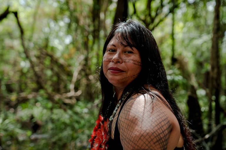 Retrato de Alessandra com a floresta ao fundo; ela é uma mulher indígena de cabelos longos e escuros, com pinturas no braço e no rosto; no pescoço usa um colar com penas vermelhas