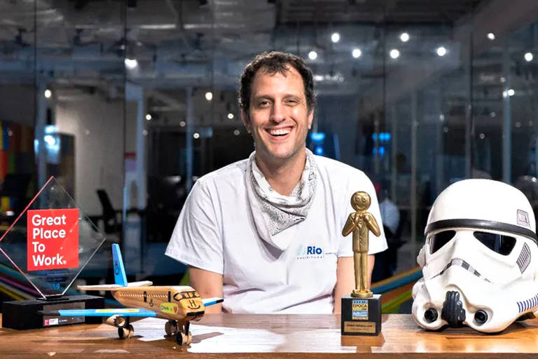 João Ricardo Mendes, CEO do Hurb, aparece sorrindo em frente a uma mesa.