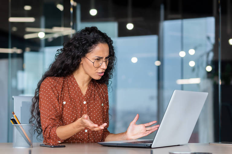 Mulher de negócios frustrada olhando para a tela do laptop, trabalhadora insatisfeita espalhando as mãos dentro do escritório, mulher latino-americana trabalhando na mesa usando computador para comunicação on-line de videochamada.
