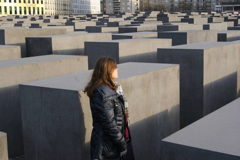 Mulher de cabelos castanhos usa casaco azul e observa o Memorial do Holocausto, em Berlim, na Alemanha, com semblante sério