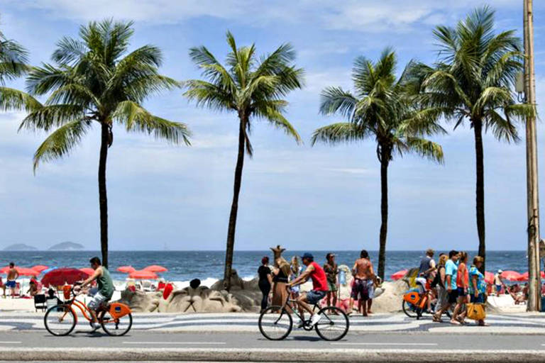 Imagem mostra calçadão de Copacabana. Na rua, estão ciclistas, na guia, pedestres, no início da praia, coqueiros, e depois areia e guarda-sóis. O mar é azul marinho e o céu limpo.