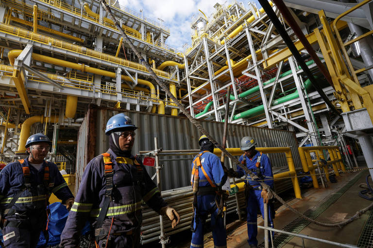 Homens com capacete e óculos de segurança caminham em navio flutuante fretado pela Petrobras. Ao fundo, estruturas de aço de cor metálica e amarelas