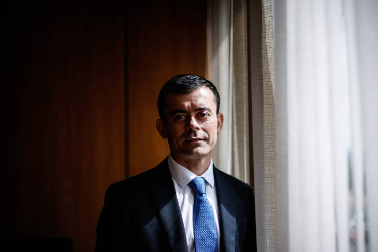 Secretário do Tesouro Nacional, Rogério Ceron, em seu gabinete no ministério da Economia. Homem branco de cabelos escuros de terno preto e gravata azul apoiado em uma janela com cortina branca. Ao fundo, uma parede de cor amadeirada 