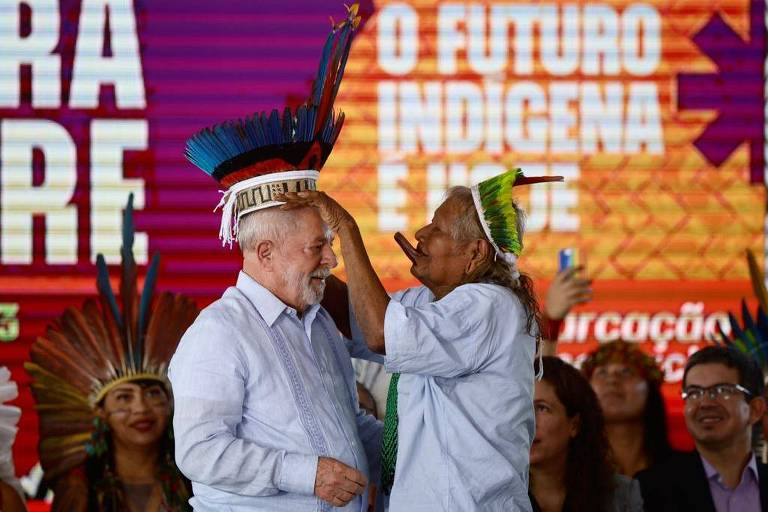 cacique raoni, à direita, coloca cocar na cabeça do presidente Lula, À esquerda. Raoni é idoso, tem cabelos grisalhos, usa cocar de penas amarelas. O cocar de lula tem penas azuis