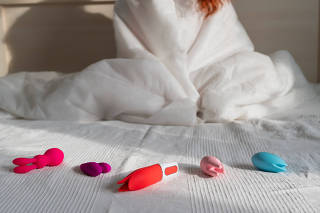 Woman sitting on bed next to a collection of vibrators and sex toys