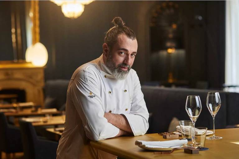 Homem branco, com barba e cabelo preso num coque, vestido de chef de cozinha, posa com os cotovelos sobre a mesa diante de um prato com duas taças