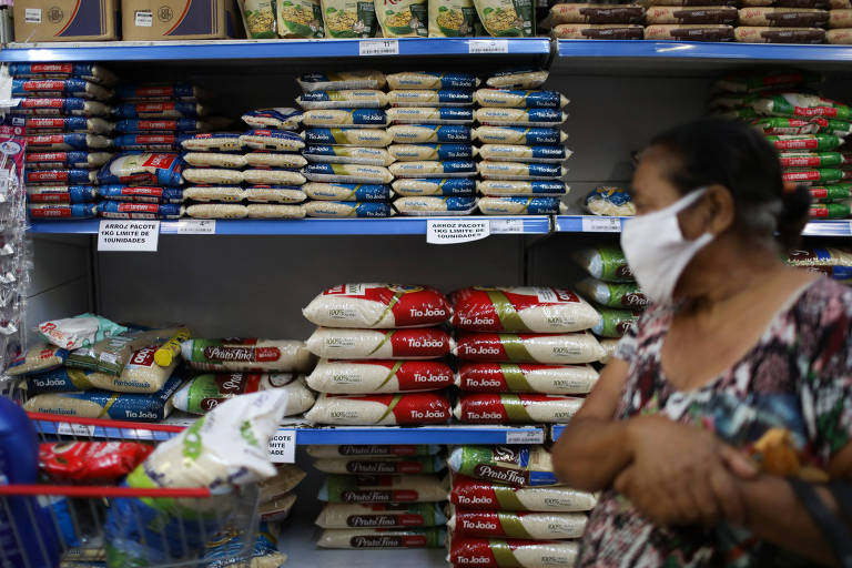 Mulher com máscara de proteção está parada ao lado de uma prateleira de supermercado repleta de sacos de arroz. Ela está com o olhar direcionado para os produtos à sua frente.