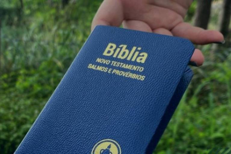 .Bíblia de capa azul com "Bíblia - Novo Testamento - Salmos e Provérbios" em letras dourados. Uma mão segura o livro e o fundo é de plantas, todo verde