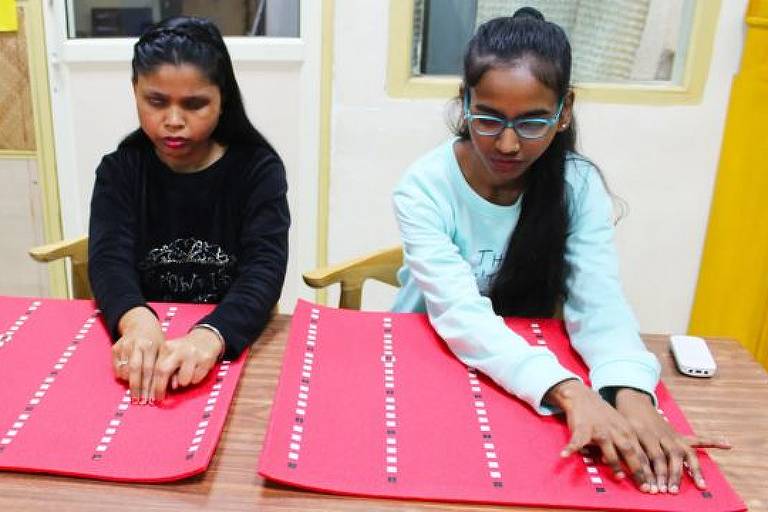 Duas jovens cegas estão sentadas lado a lado de frente para uma mesa. Sobre a mesa há painéis de treinamento que ambas tateiam no treinamento para identificação de anomalias na mama