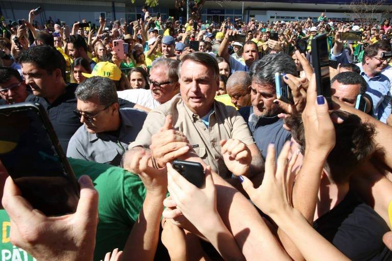 Bolsonaro, um homem branco, cabelos castanhos e blusa bege, cumprimenta várias pessoas que estendem os braços em sua direção, alguns com celulares apontados. Ele caminha cercado por homens no meio de uma grande aglomeração