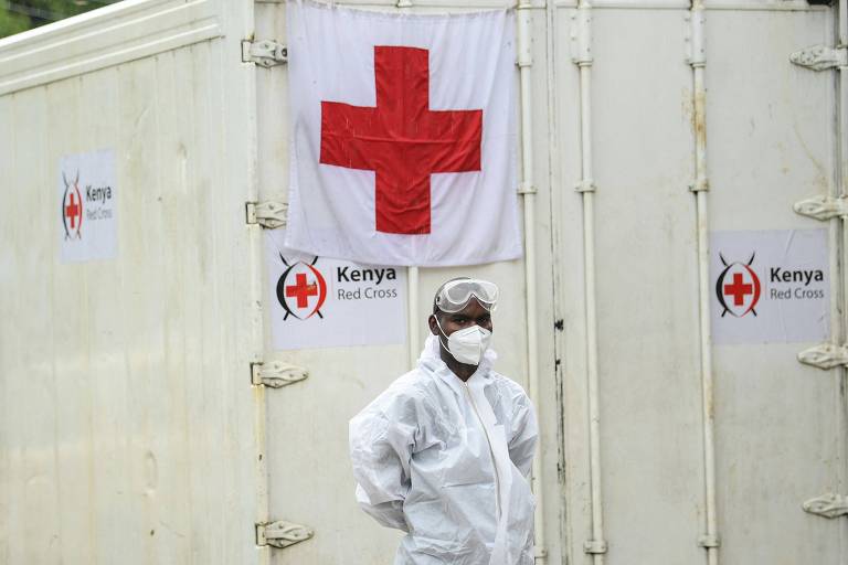 Homem negro vestindo roupas brancas características de um profissional da saúde está de pé à frente de uma porta branca onde está pendurada uma bandeira também branca com uma cruz vermelha.
